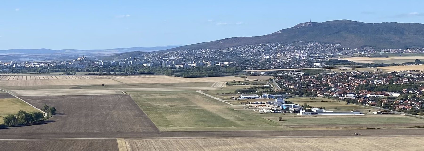 Letisko Nitra z výšky — dve rovnobežné trávnaté dráhy, hangáre,
              za nimi Janíkovce, Nitra a Zobor